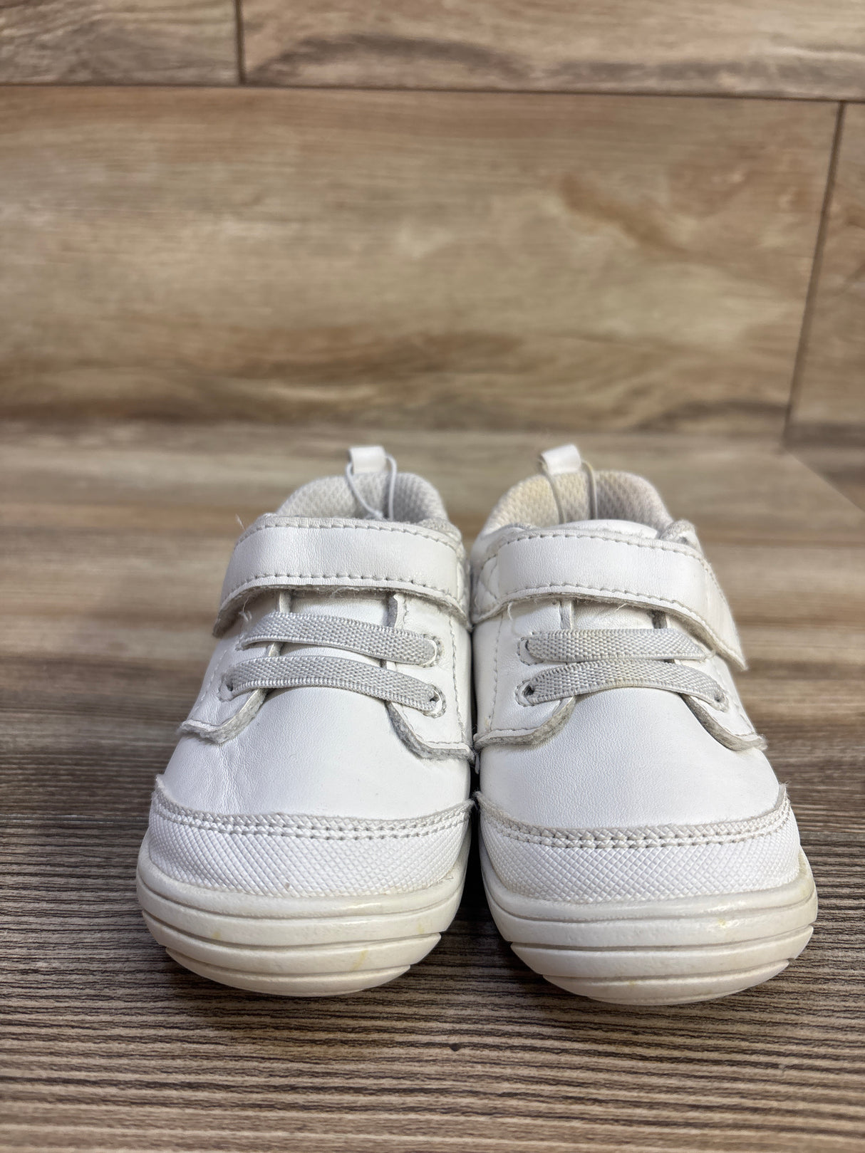 Front view of Stride Rite Taye 2.0 white children’s sneakers showcasing hook-and-loop straps and textured soles on a wood floor