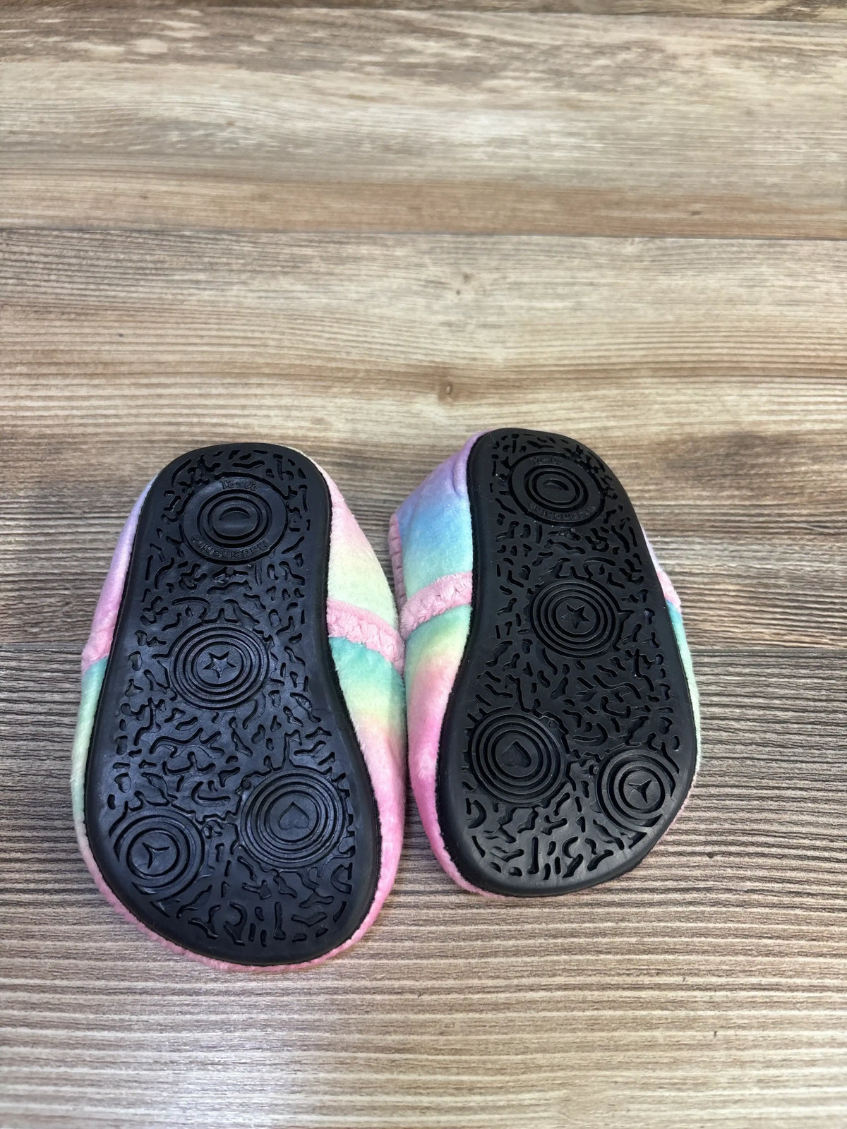 Bottom view of unicorn toddler slippers size 5C showing black textured rubber soles and pastel rainbow plush uppers on wood floor