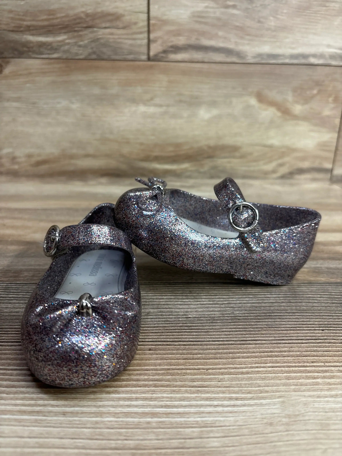 Angled front of Mini Melissa Sweet Love jelly flats size 7C with silver glitter bow and Mary Jane buckle on wood surface