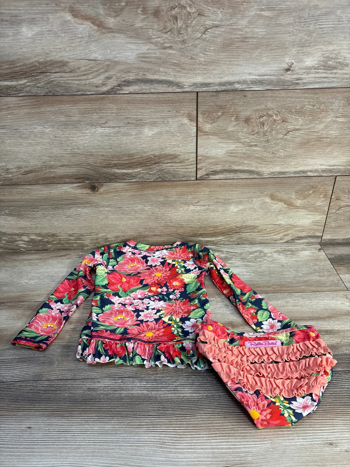 Flat lay of Ruffle Butts navy floral rashguard set with ruffle-hem long-sleeve top and coral ruffle bottoms on wood floor