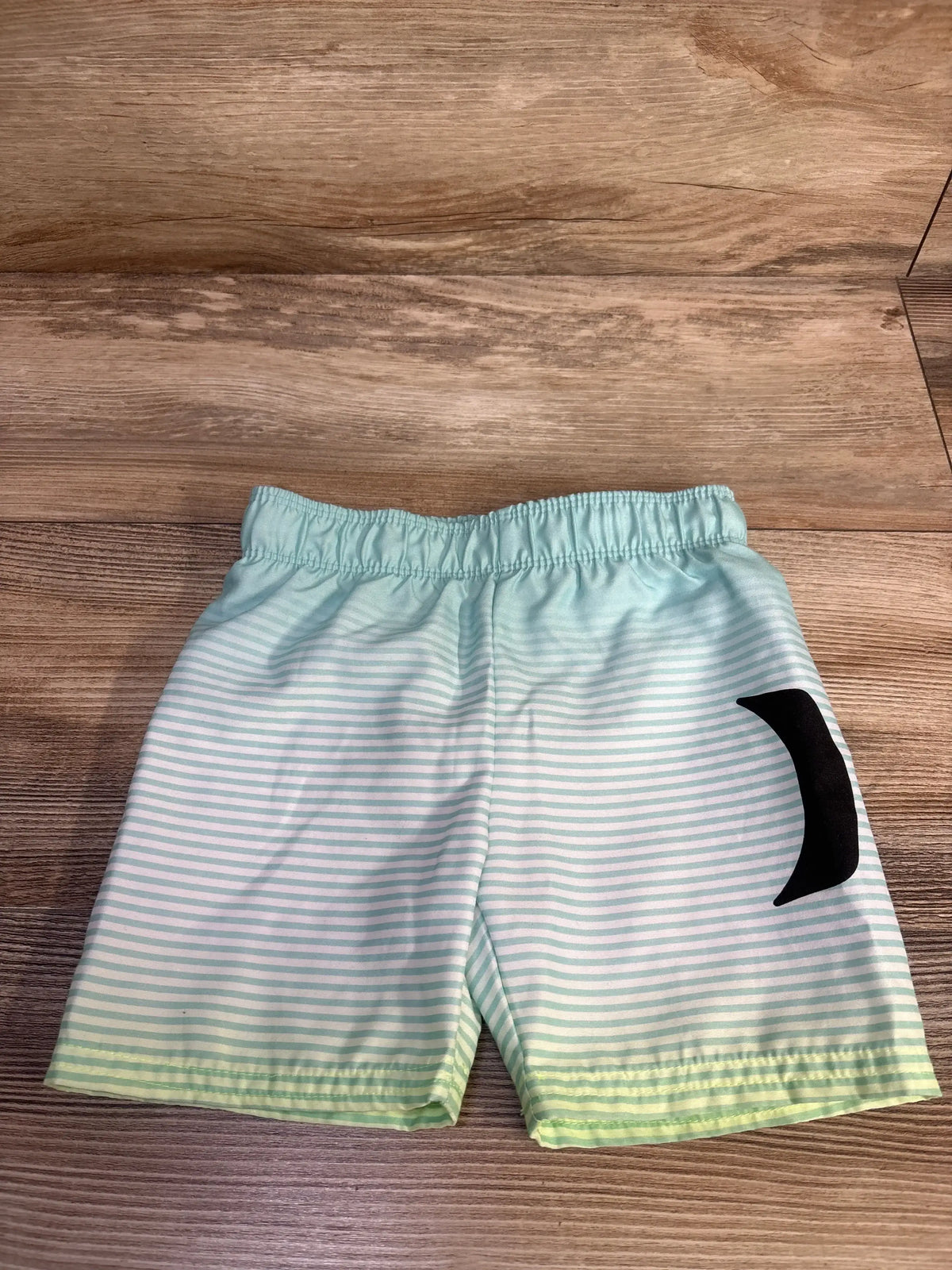Front view of boys' Hurley swim trunks with light blue-and-white stripes, elastic waist, and black logo on wood background