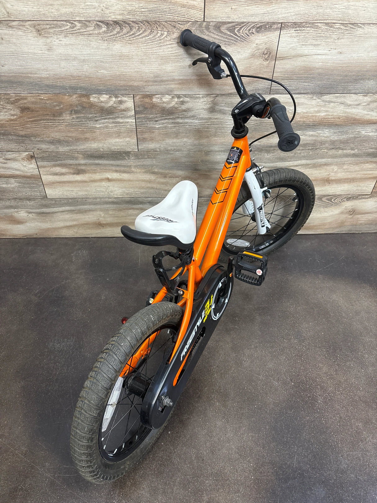 Orange RoyalBaby Freestyle kids bike with white seat, black handlebars, and training wheels on wood floor