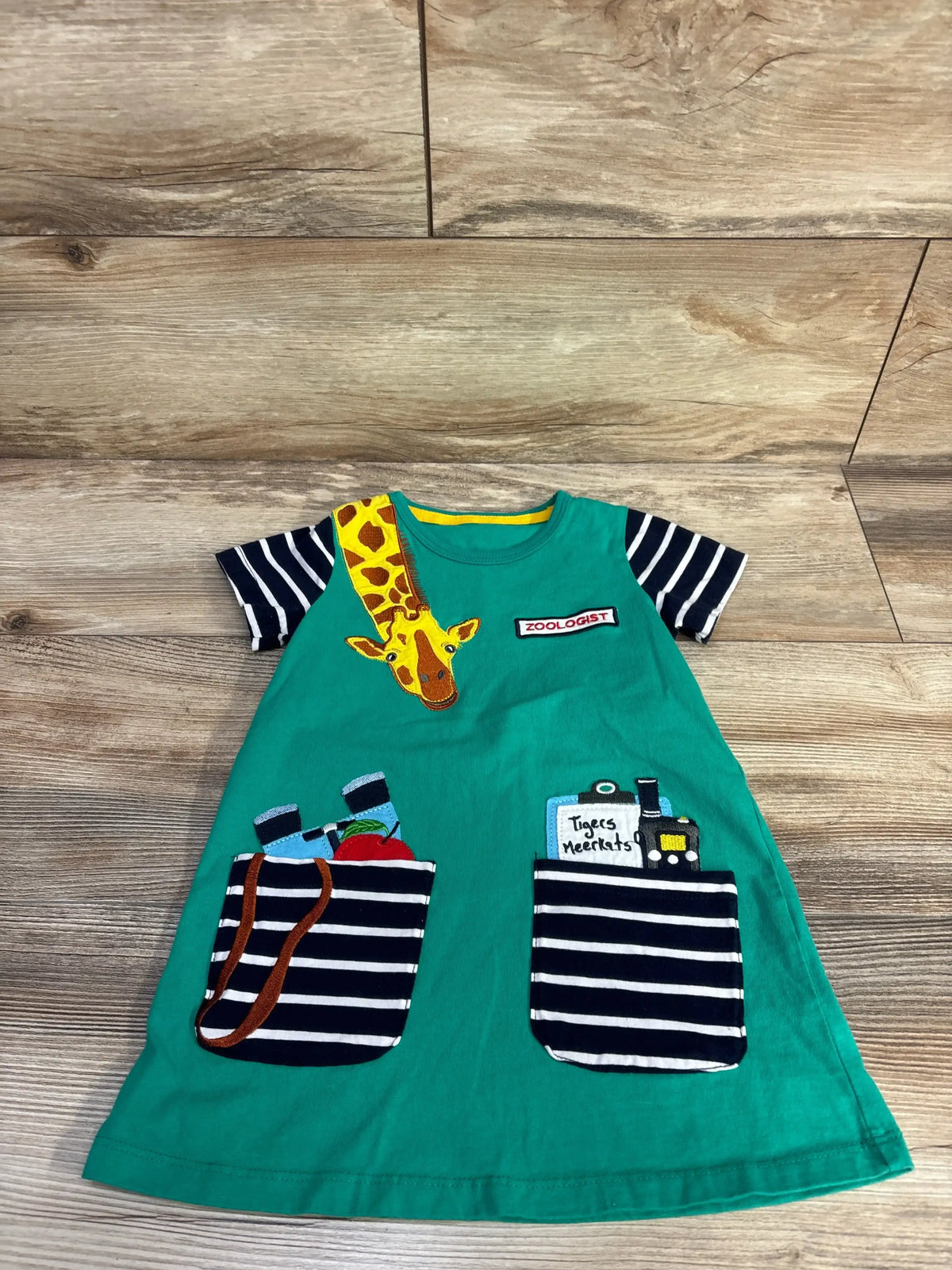 Flat lay of green toddler T-shirt dress showcasing giraffe applique, navy-striped pockets and sleeves and ZOOLOGIST patch with wood floor background