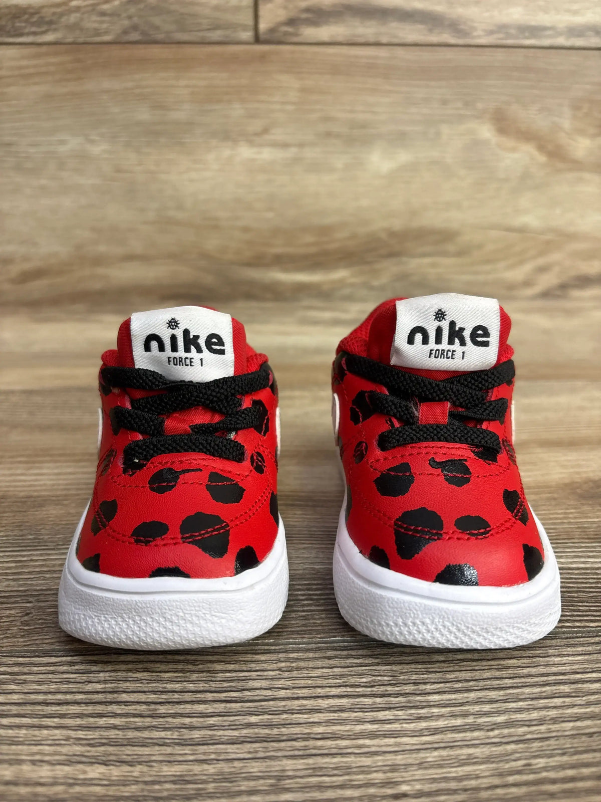 Pair of red Nike Force 1 kids sneakers with black spots and black laces on wood floor