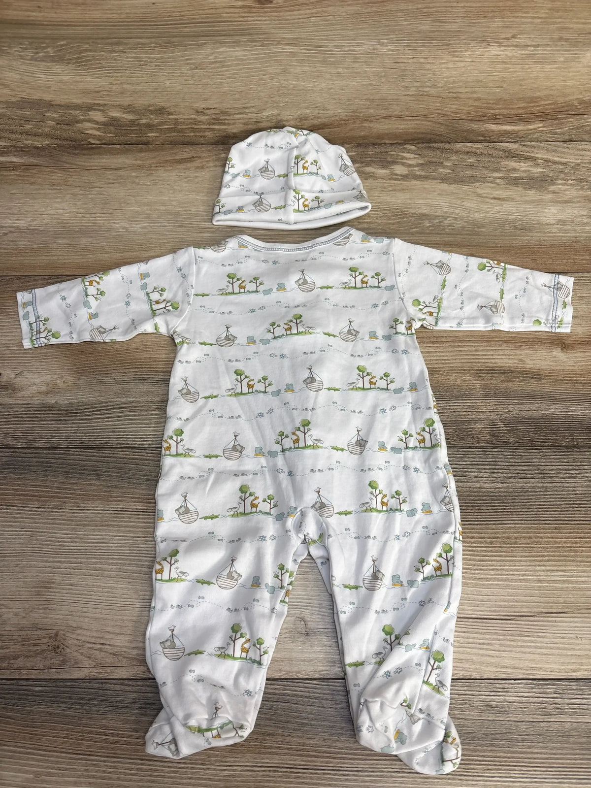 Top-down view of white 2-piece footed baby sleeper and matching hat with Noah-print boats and trees on wooden background
