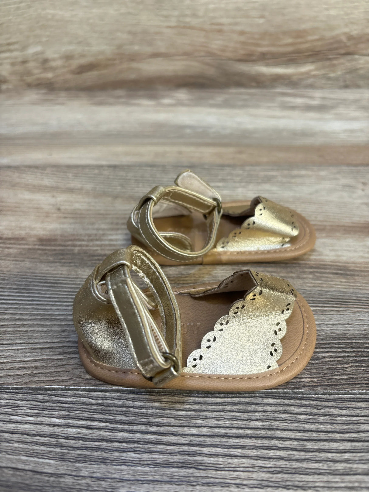 Gold baby sandals with ankle straps and scalloped edges on wood background