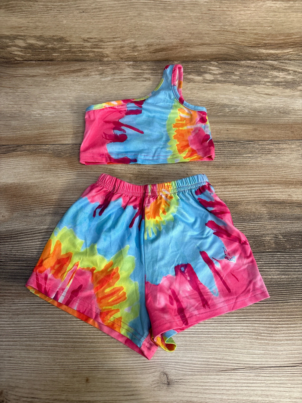 Flat lay of multicolor tie-dye baby tank and shorts set with one-shoulder top and elastic waist on light wood background
