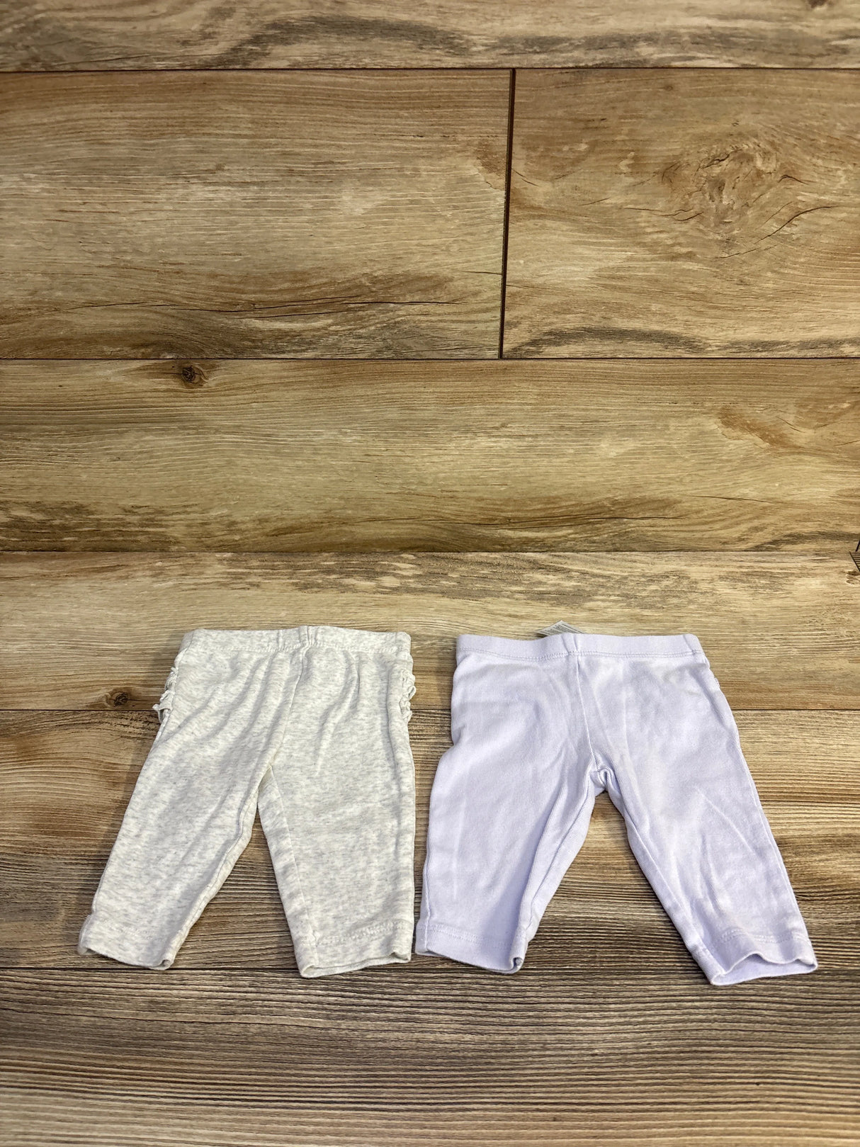 Flat lay of two Carter’s baby ruffle pants, purple and heather gray, showing elastic waistbands and side ruffles on wood background
