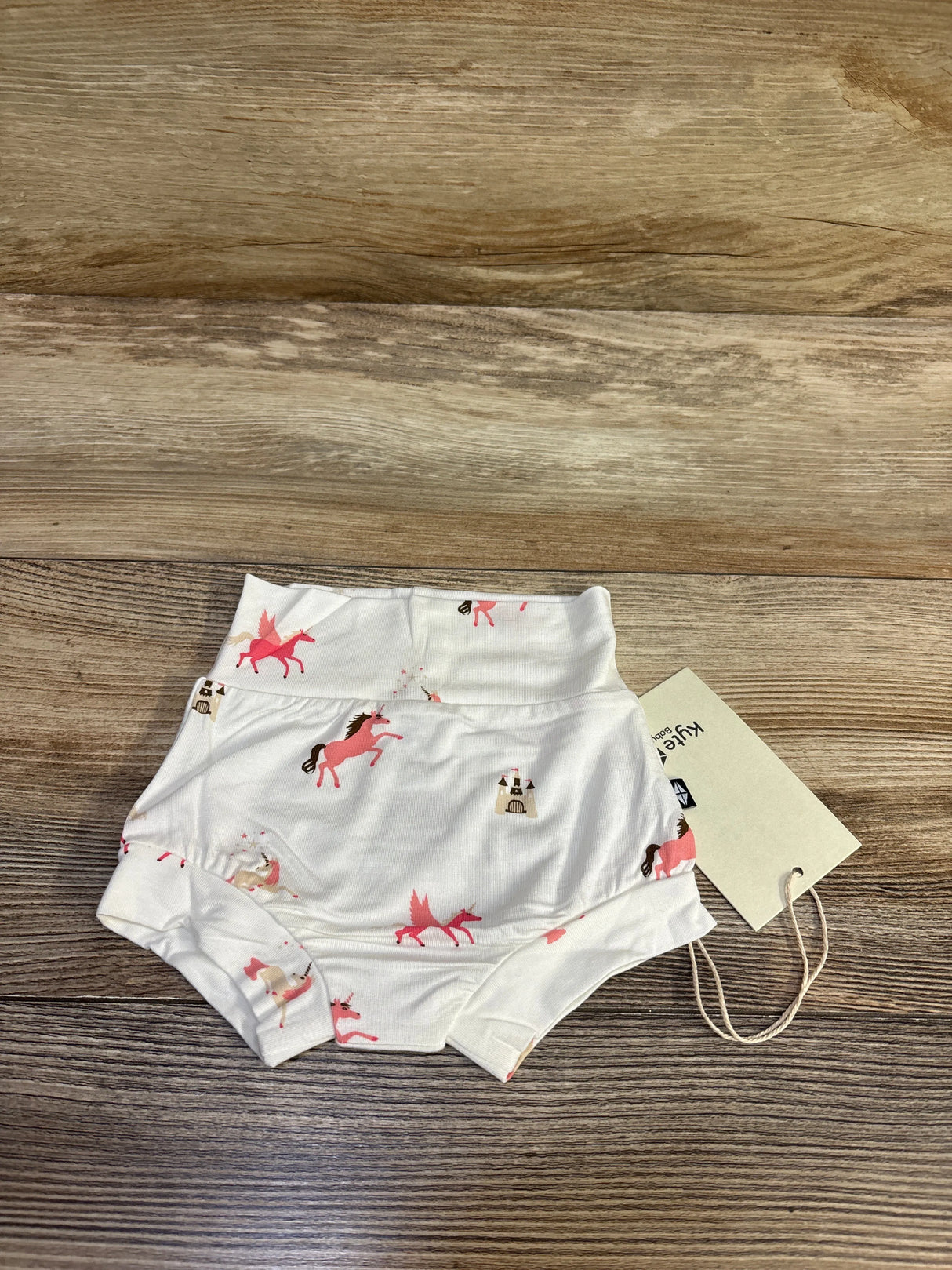 Top-down view of white Kyte Baby bamboo bummie with pink unicorn and castle print, high-waisted style on wooden floor