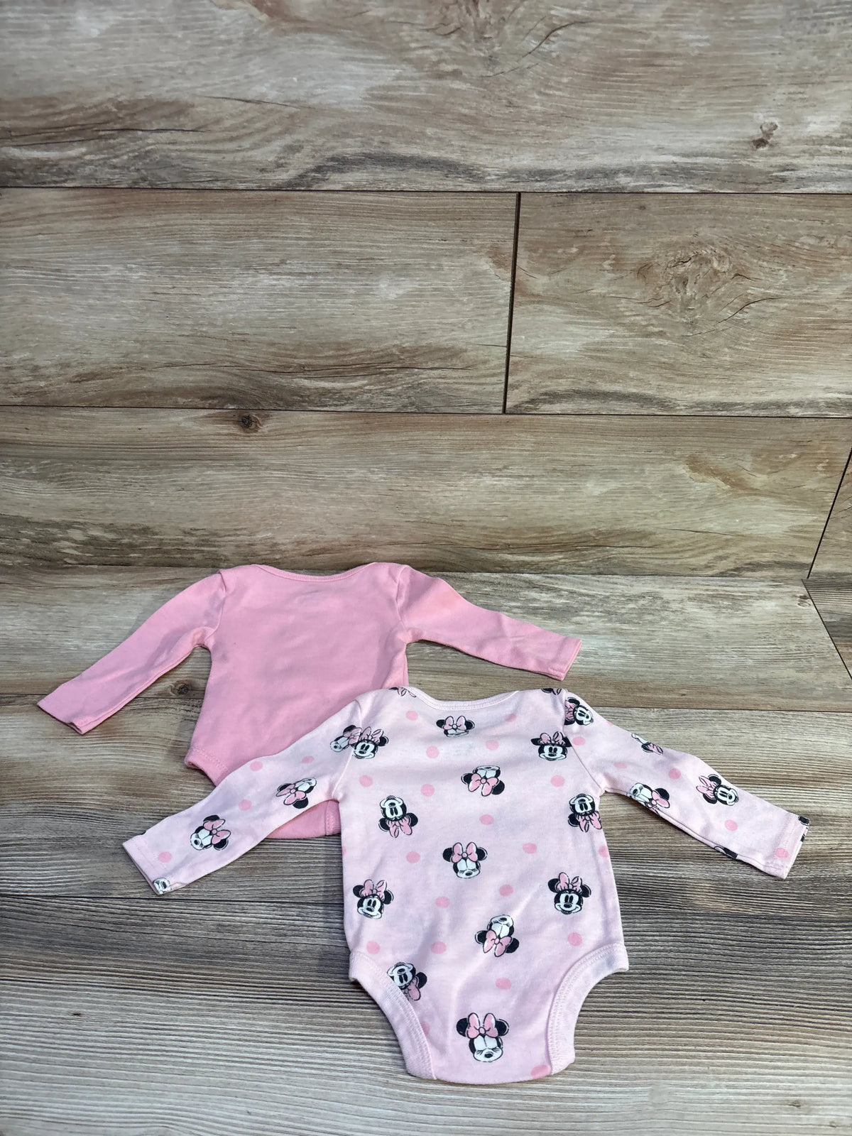 Top view of Disney Baby two-pack long-sleeve Minnie Mouse bodysuits in pink, one solid and one Minnie Mouse print, on a wood-look floor