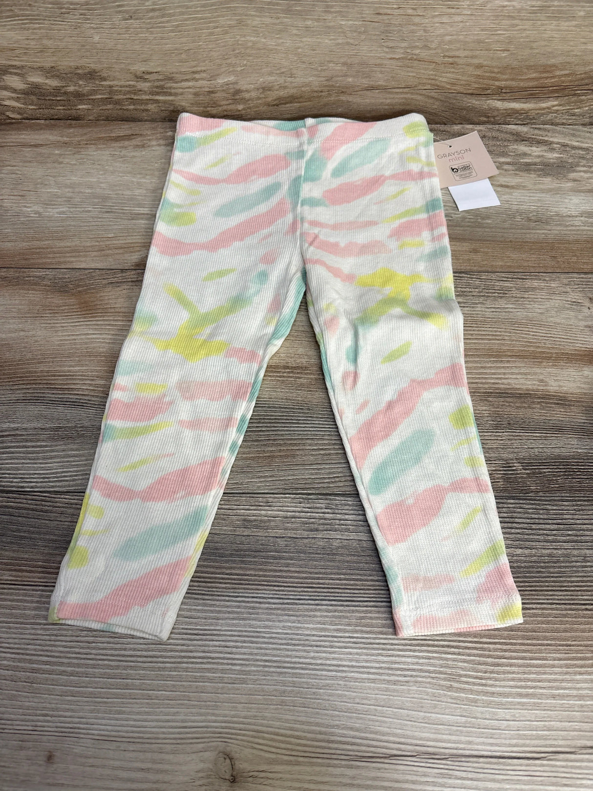 Flat lay of Grayson Mini tie-dye toddler leggings, cream ribbed with pastel pink, mint and yellow stripes on wood