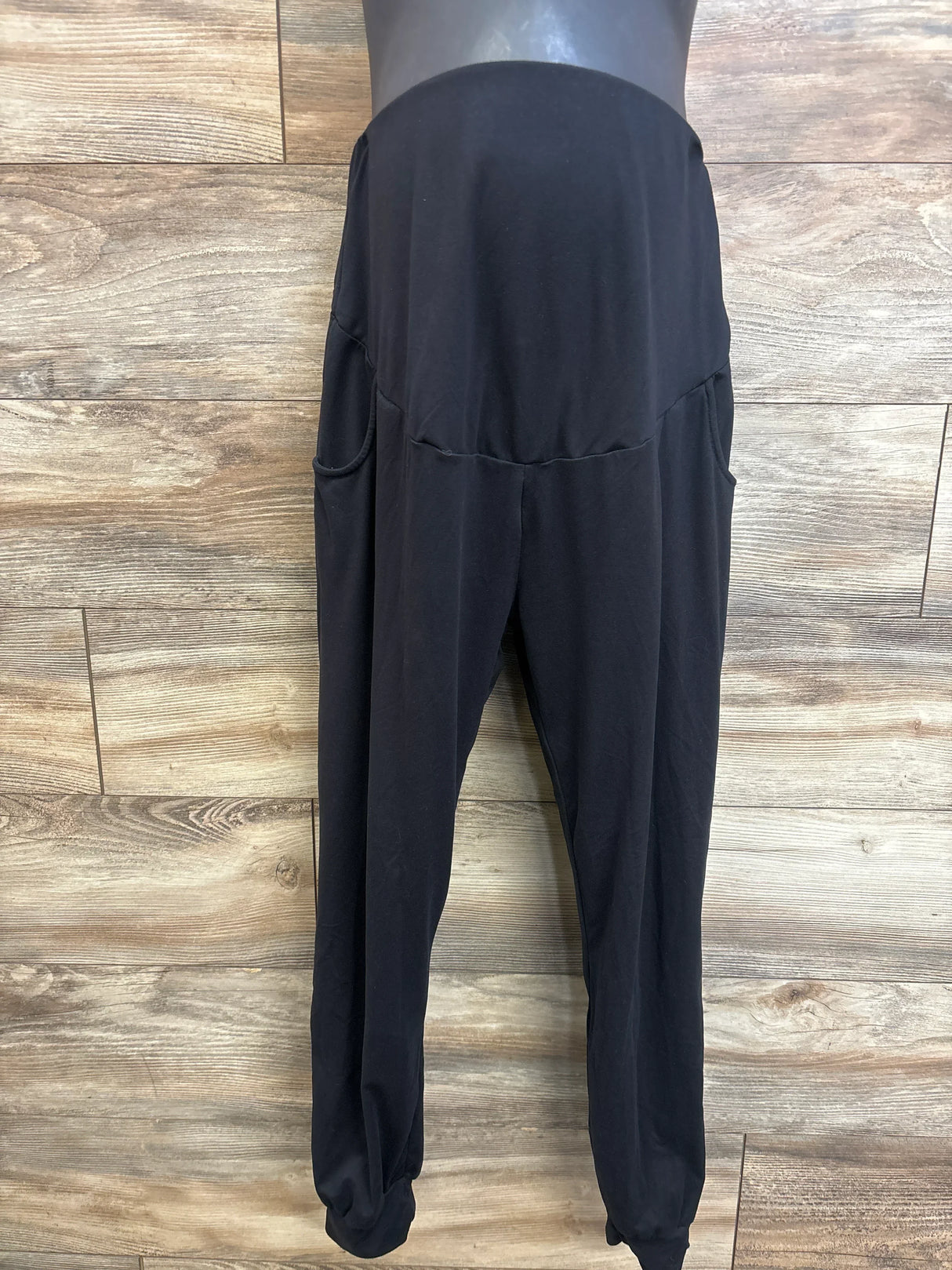 Front view of black maternity leggings with over-bump belly panel, side pockets, and tapered cuffed ankles on a light wood background