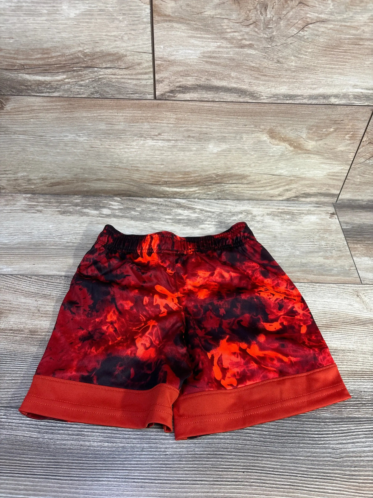 Flat lay of red and black tie-dye Under Armour toddler shorts (4T) showing elastic waistband and cuffed hem on wood floor