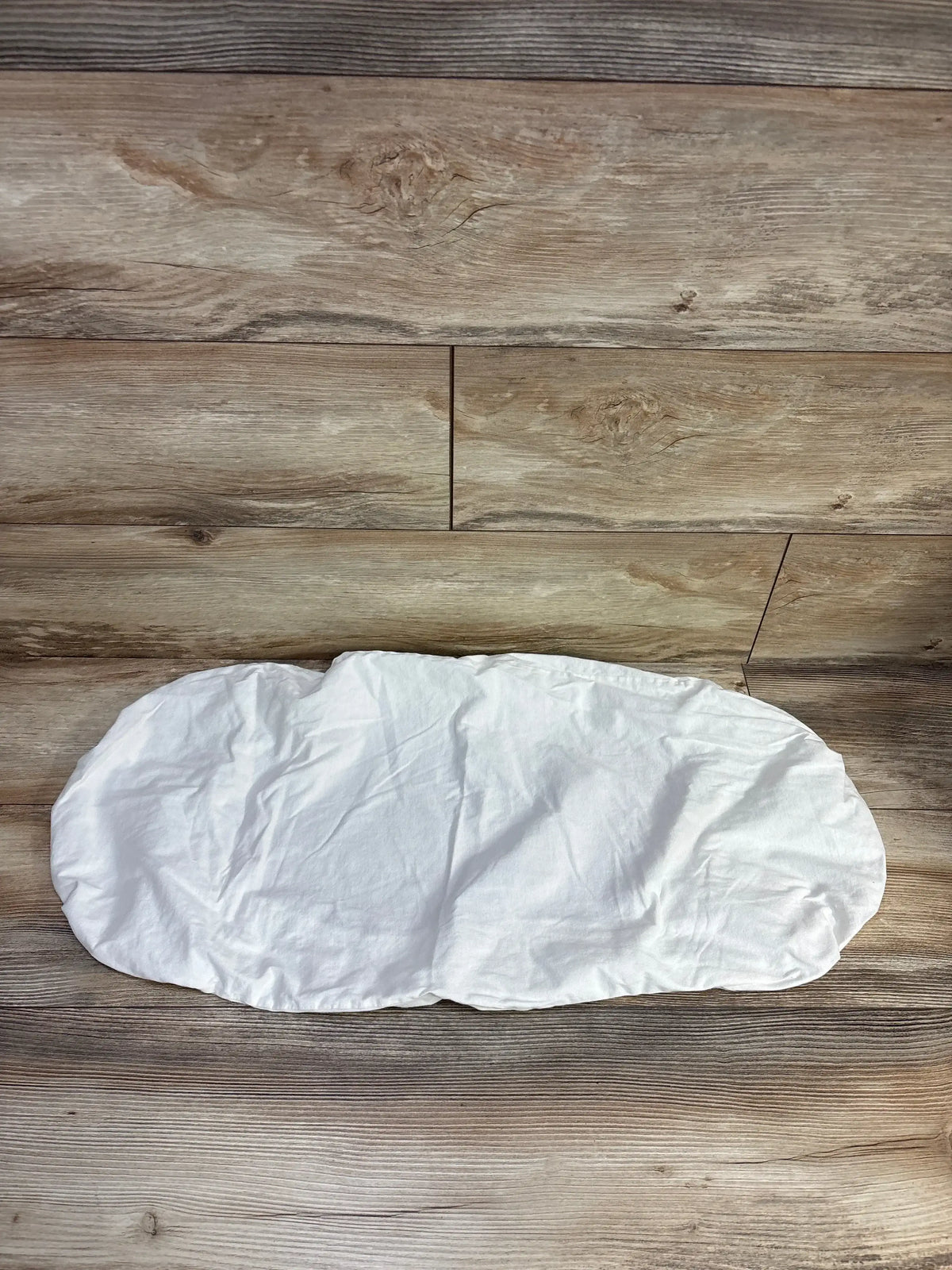 Top-down view of Happiest Baby SNOO bassinet sheet in white showing contoured oval shape and elasticized edges on wood floor background