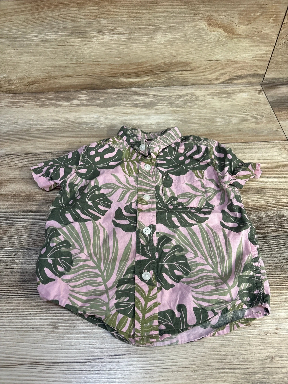 Front flat-lay of toddler button-up shirt in pink with green tropical leaf print, chest pocket and white buttons on wood