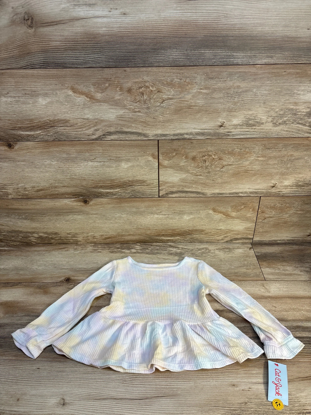 Long sleeve pastel tie-dye toddler shirt with ruffled hem, laid flat on wood floor.