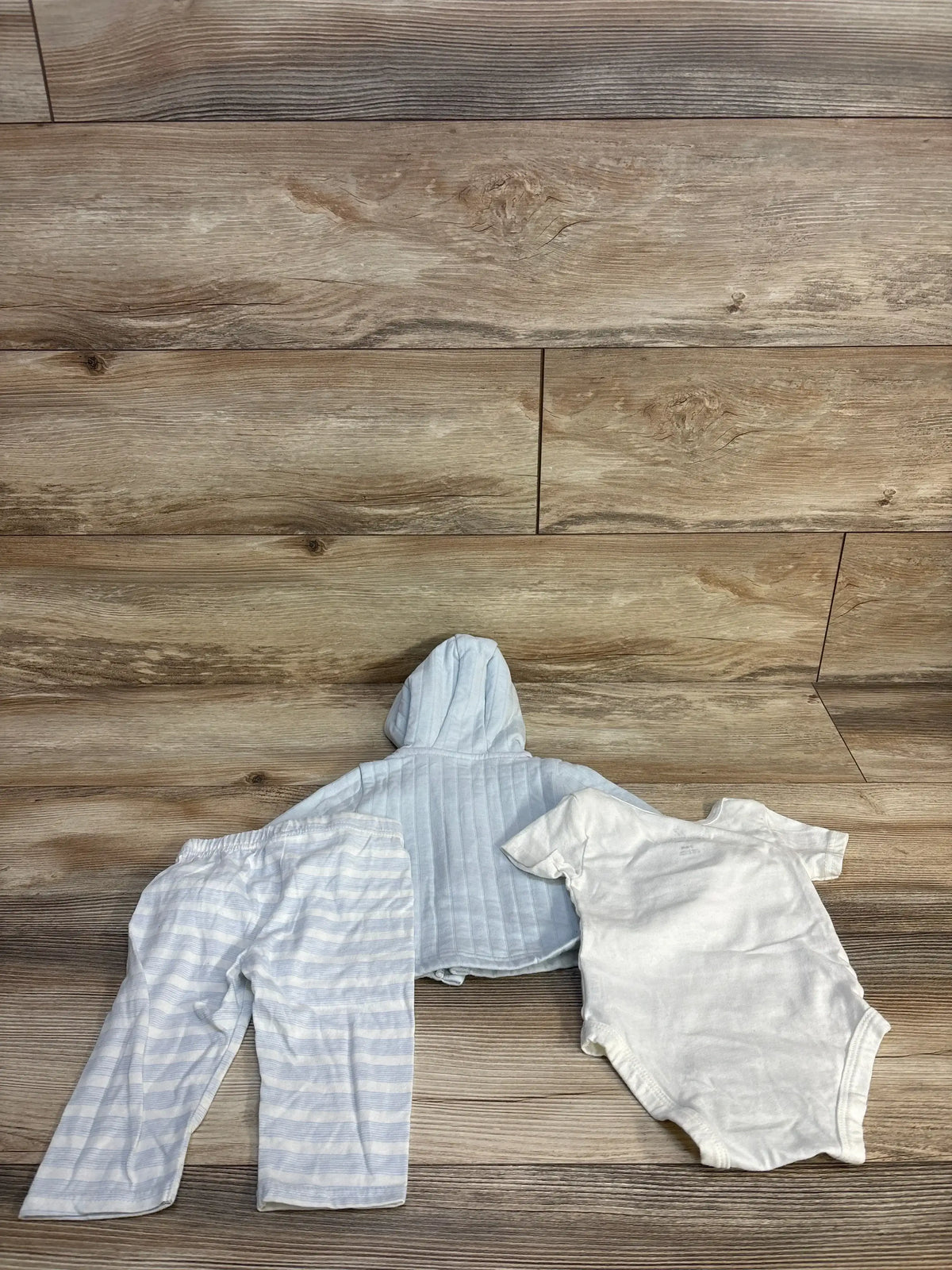 Flat lay of Peter Rabbit 3-piece baby outfit: blue hooded jacket, striped pants, and white bodysuit on wood floor