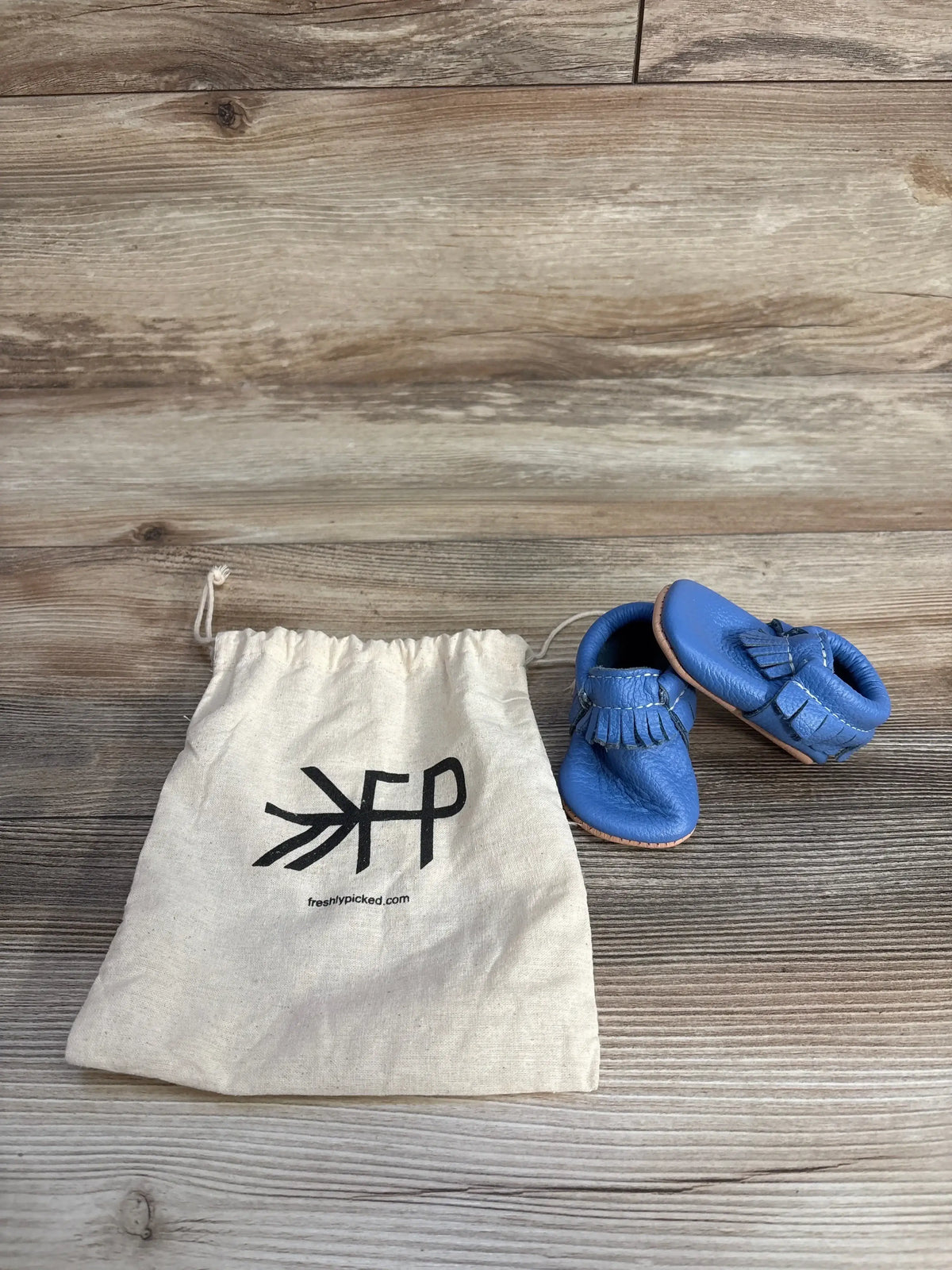 Blue baby moccasins with fringe next to Freshly Picked cloth bag on wood background