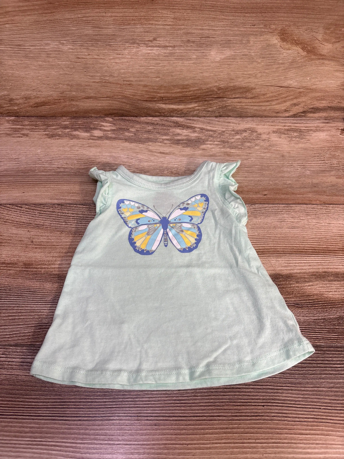 Front view of First Impressions butterfly tank top showcasing teal fabric and butterfly graphic with ruffled straps on a wood background