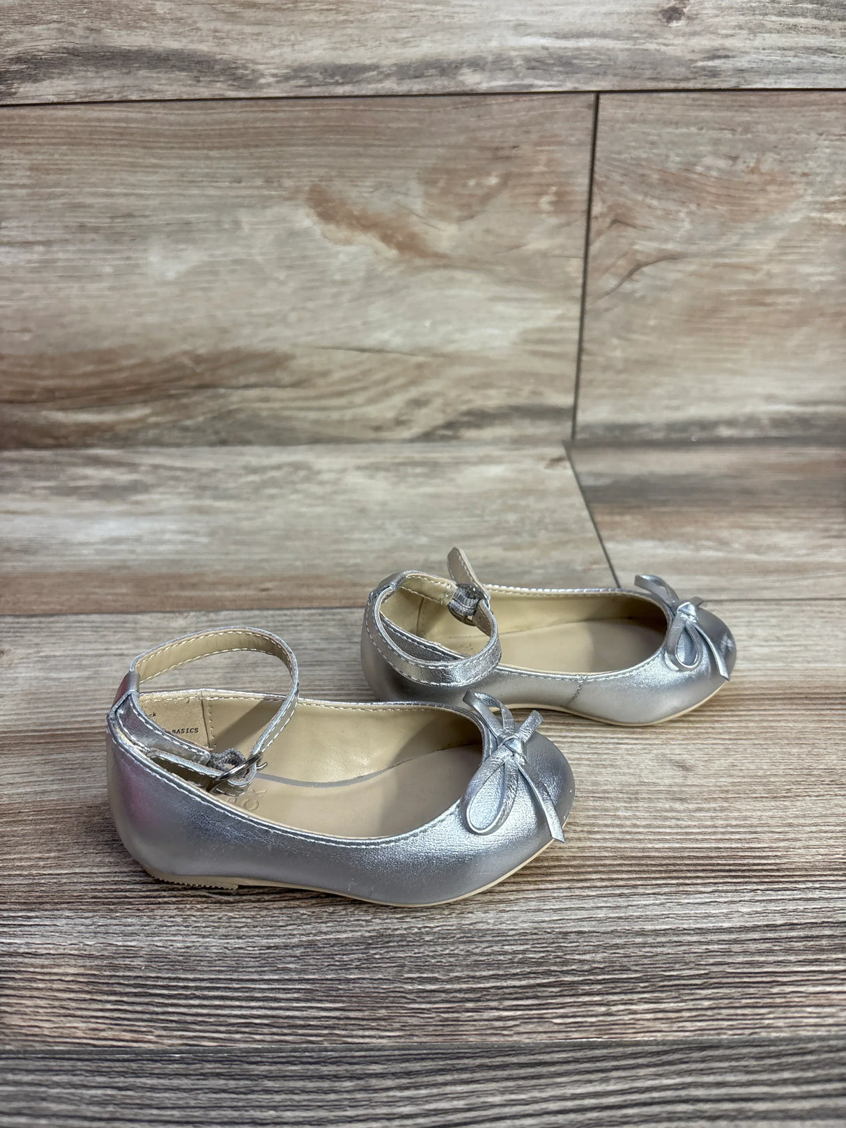 Silver toddler girls' ballet flats with ankle straps and bow detail on wooden floor