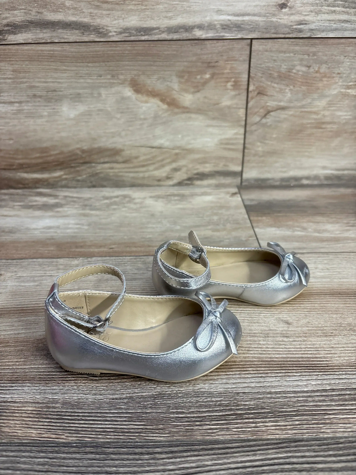 Silver toddler girls' ballet flats with ankle straps and bow detail on wooden floor