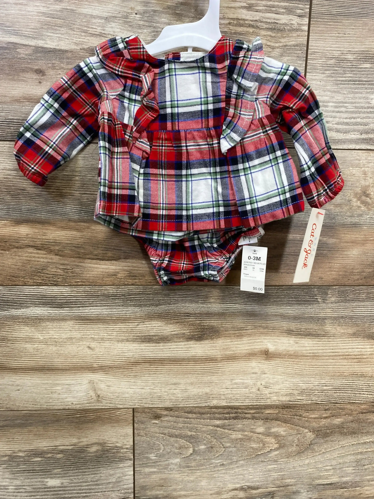 Flat-lay of Cat & Jack 2-piece red plaid baby top and bottoms with ruffle collar and sleeves on wood floor, 0-3M tag