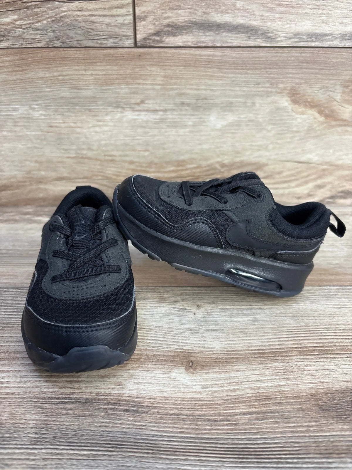 Front three-quarter view of black Nike Air Max Motif PS toddler sneakers showing visible Air Max heel unit and mesh upper on wood plank background