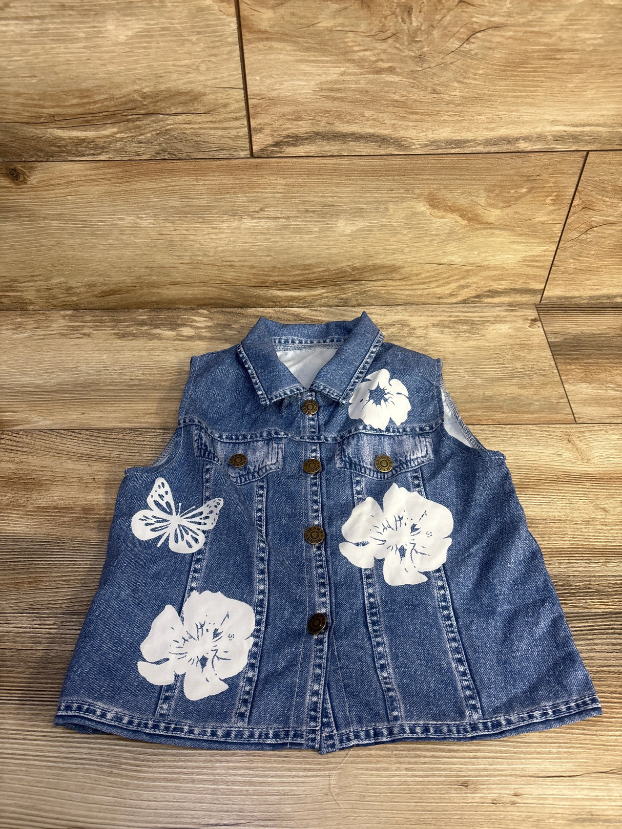 Flat lay of denim-look sleeveless button-up shirt size 4/5T showing white floral and butterfly prints and metal buttons