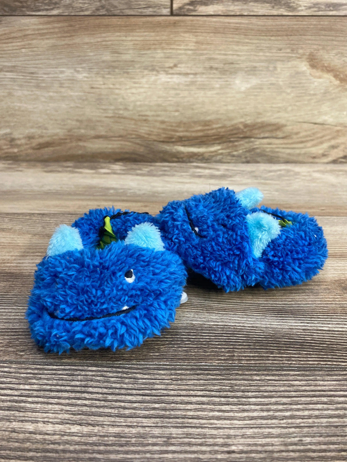Pair of blue fuzzy baby slippers with smiling monster faces and light blue horns on a wooden surface