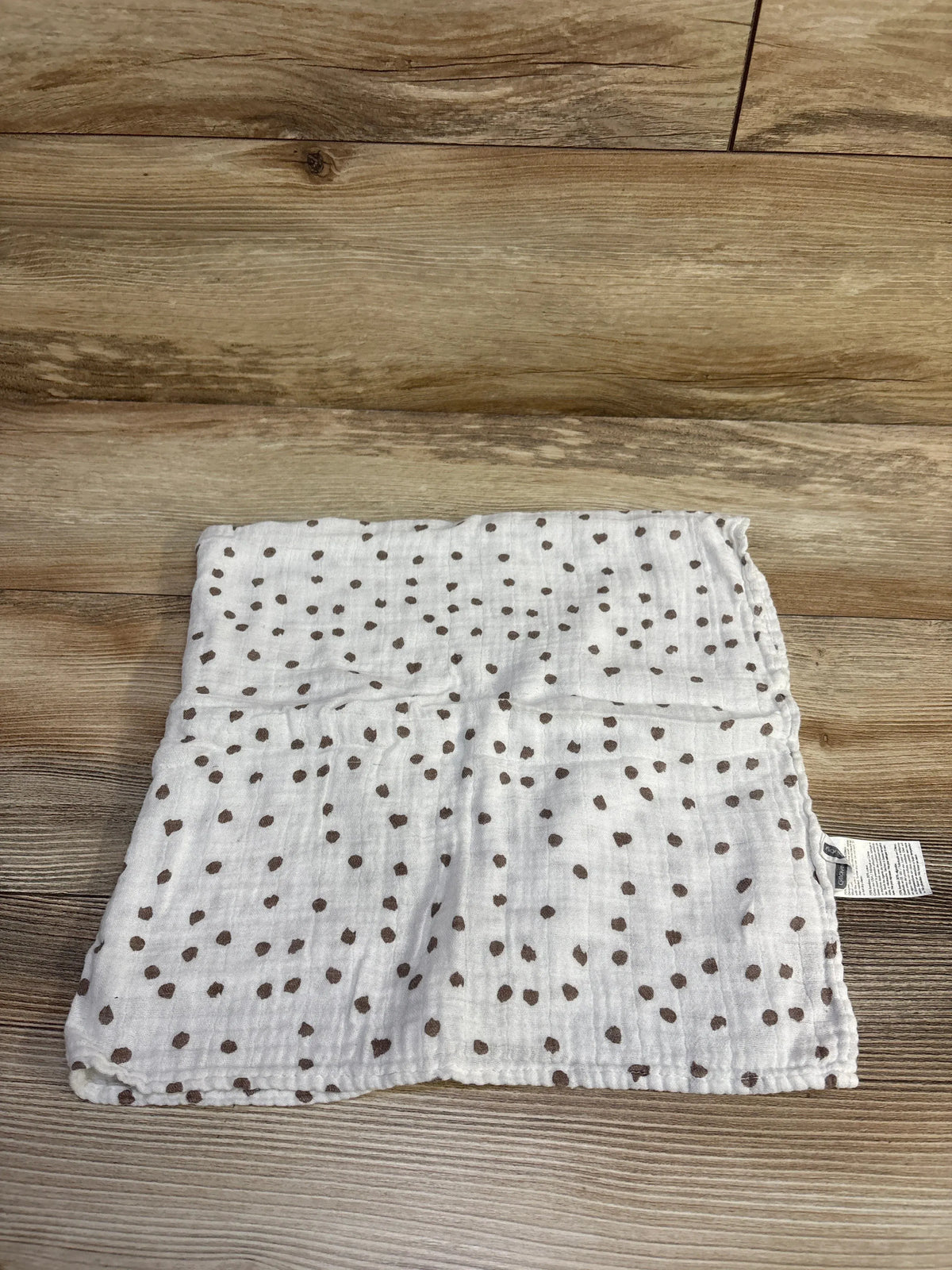 Top-down view of white muslin-bamboo swaddle blanket 28 x 28 in showing scattered brown dot print on wooden floor