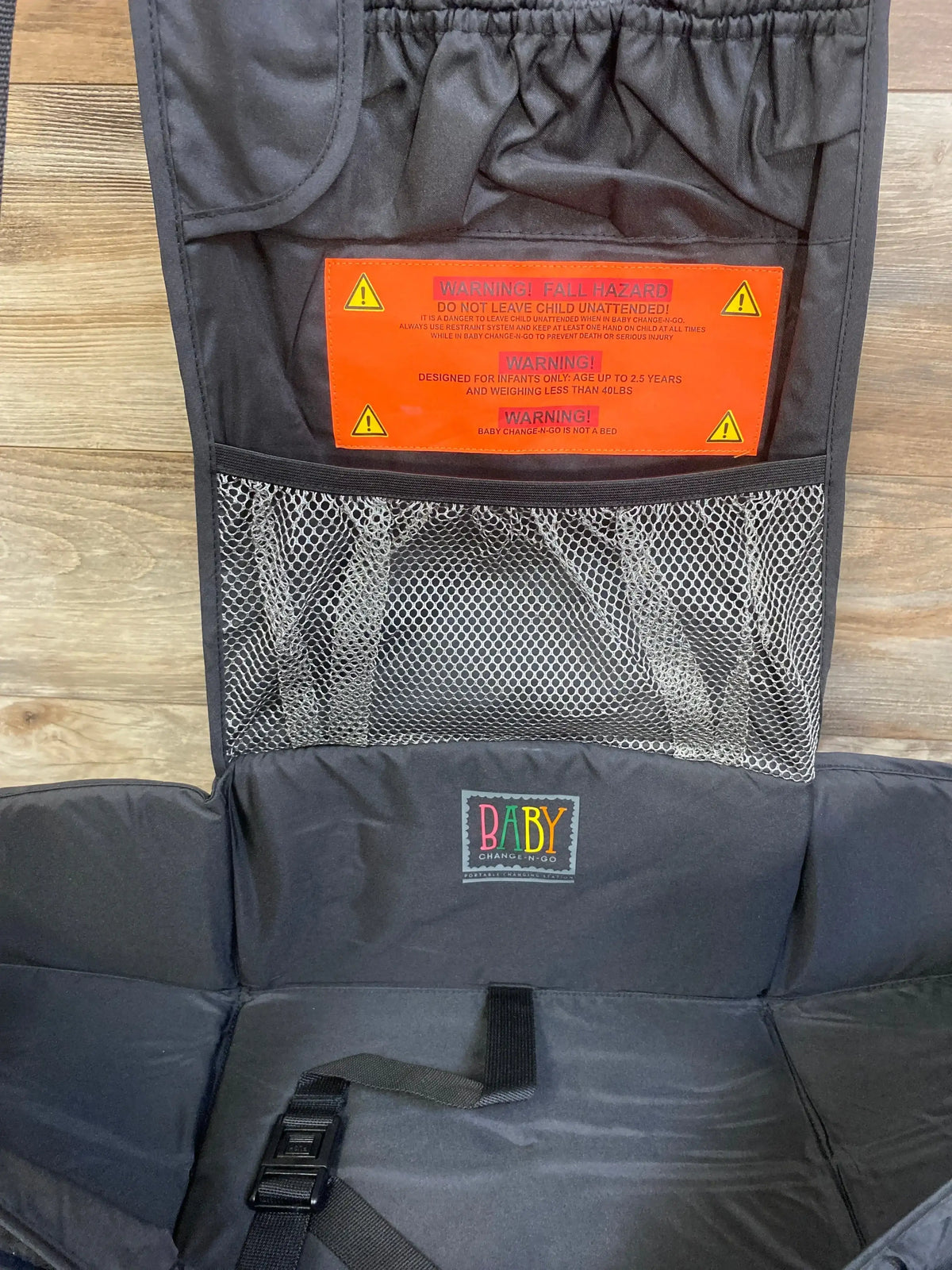 Front of Baby Change-N-Go hanging changing station with mesh pocket, safety strap, padded pad and orange label reading WARNING
