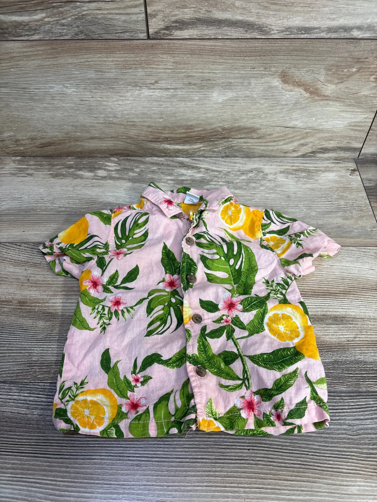 Flat lay of pink linen-blend toddler button-up shirt showing green leaves, pink flowers, and yellow citrus on wood background