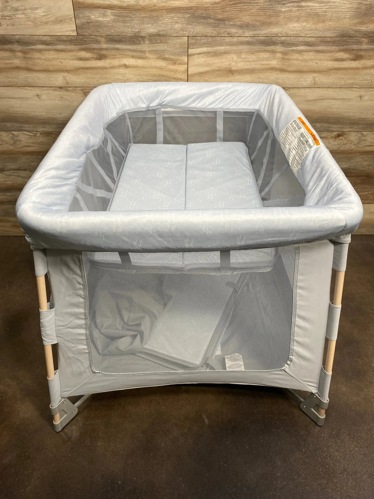 Top-front view of Maxi-Cosi Swift playard in classic slate showing mesh sides, mattress, and wood legs against wood-paneled background