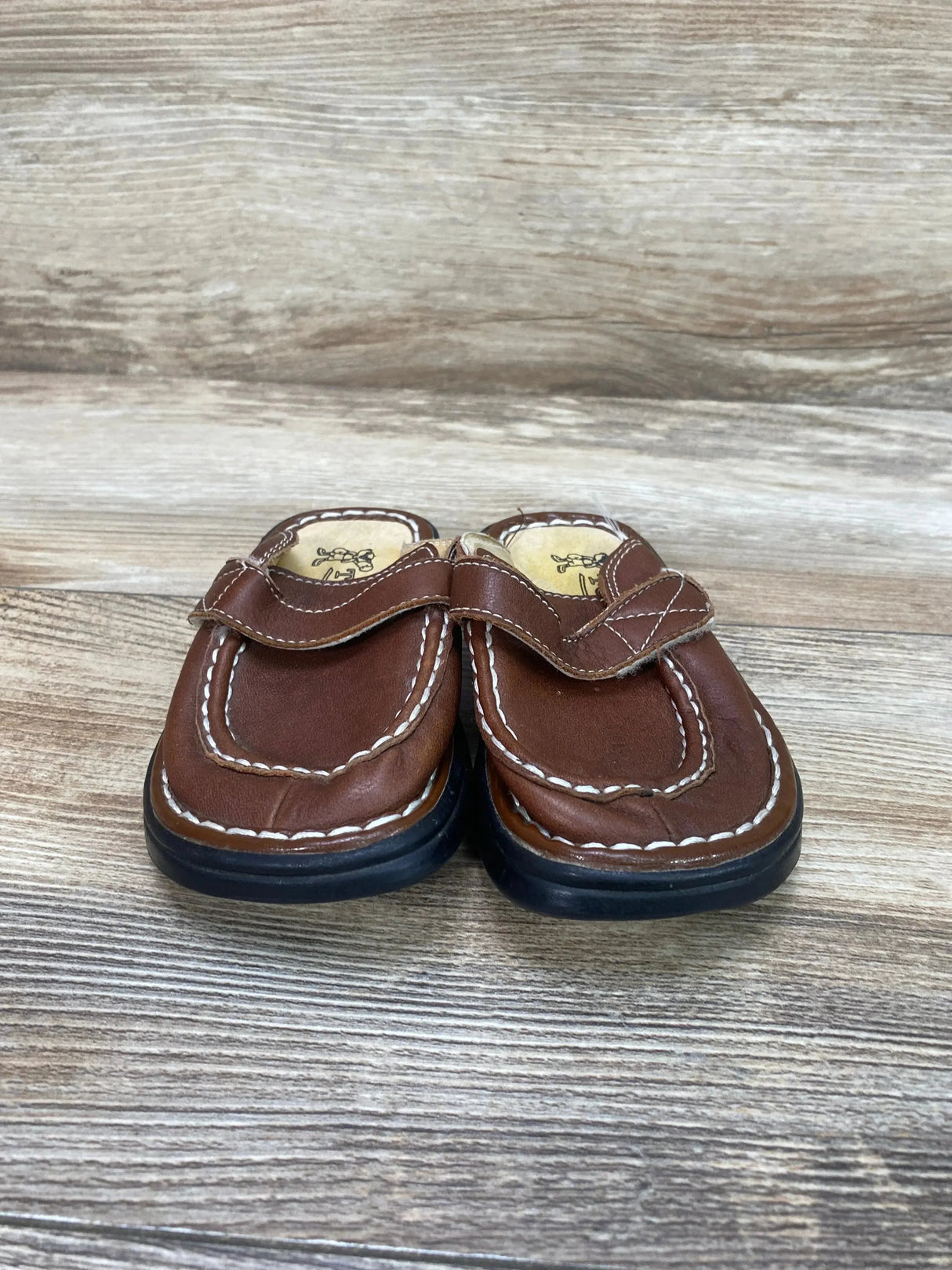 Front view of Tom Dog brown leather clogs size 5/6C with white stitching and hook-and-loop strap on wood background