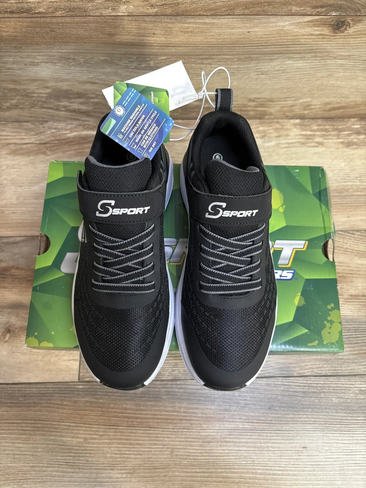 Black S Sport athletic sneakers on green shoe box, wood floor background