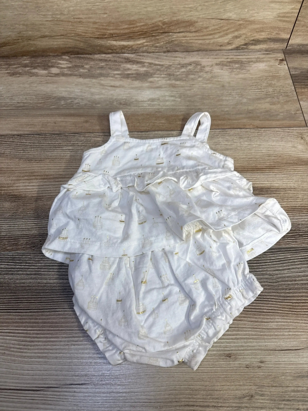 Flat lay of Baby Gap white two-piece birthday cake outfit, ruffled top and bloomers with gold cake print on wood floor
