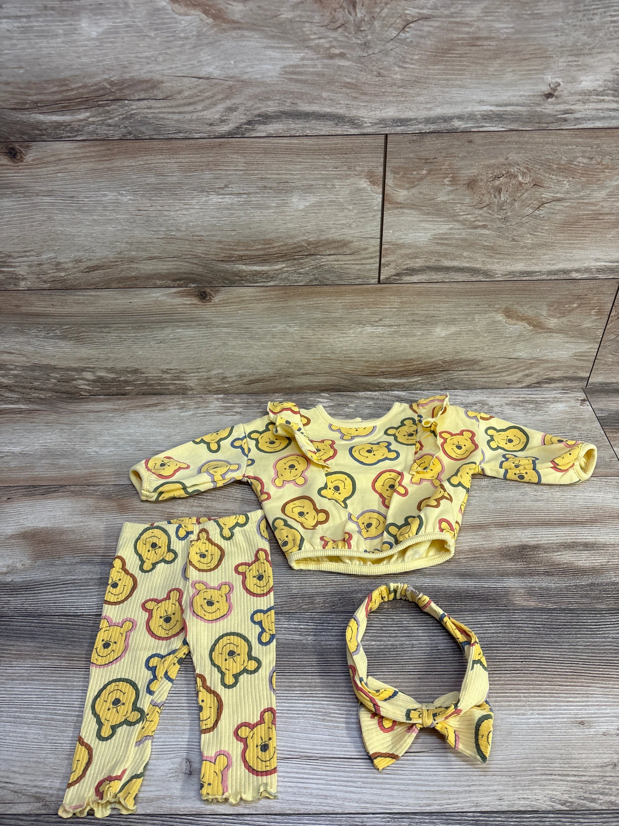 Flat lay of yellow Disney Baby 3-piece Winnie the Pooh set, sweatshirt, leggings, and headband on wood background, size 3-6M