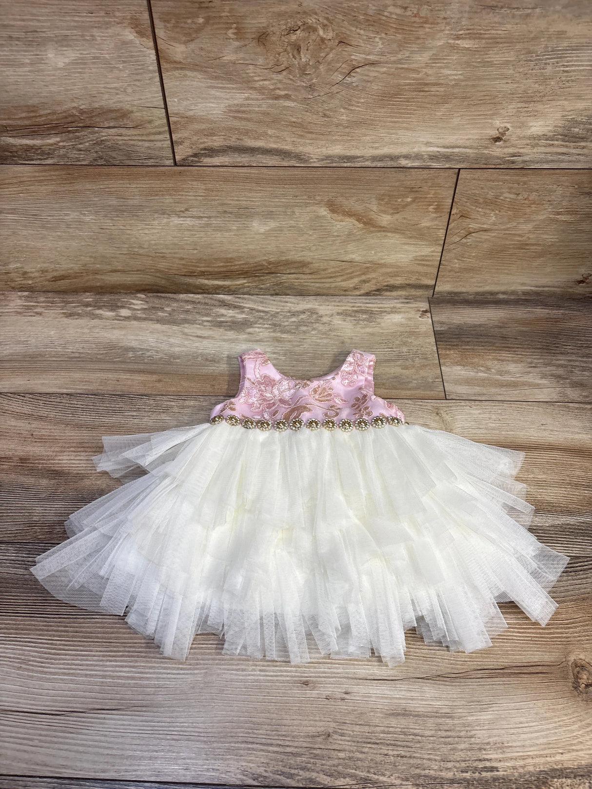 Flat lay of infant sleeveless dress and bloomers showing pink floral bodice, gold beaded trim, layered white tulle skirt on wood background