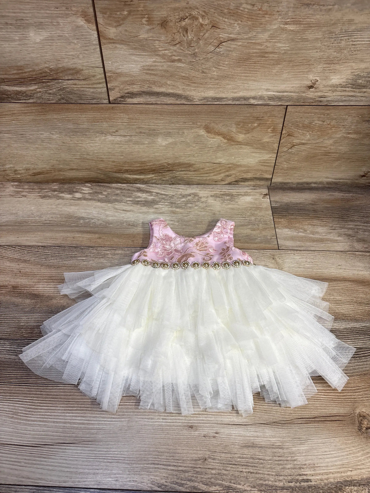Flat lay of infant sleeveless dress and bloomers showing pink floral bodice, gold beaded trim, layered white tulle skirt on wood background