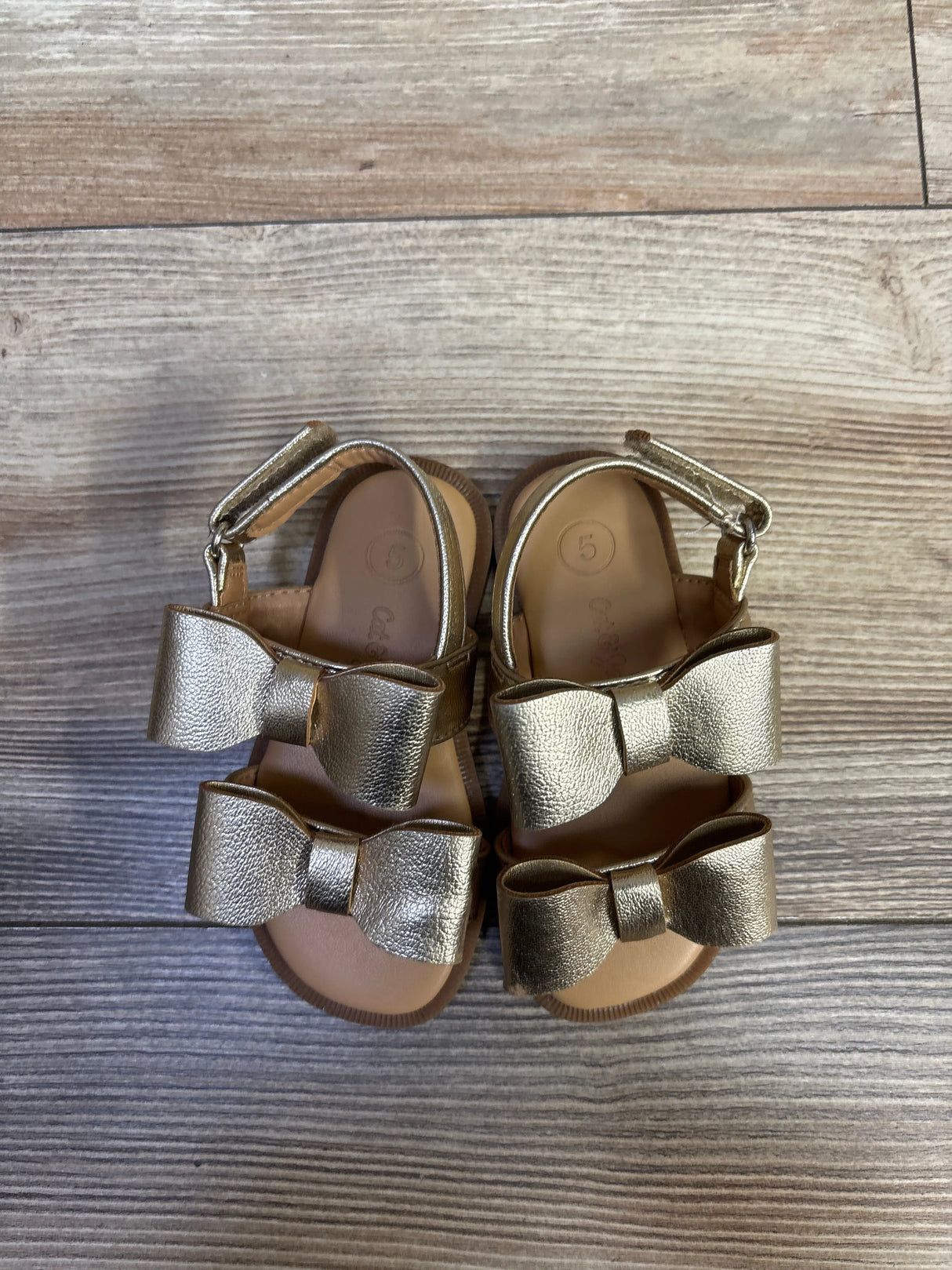 Top view of Cat & Jack Ruby toddler gold bow sandals showing two metallic bows, ankle strap, and size 5 insole on wood floor