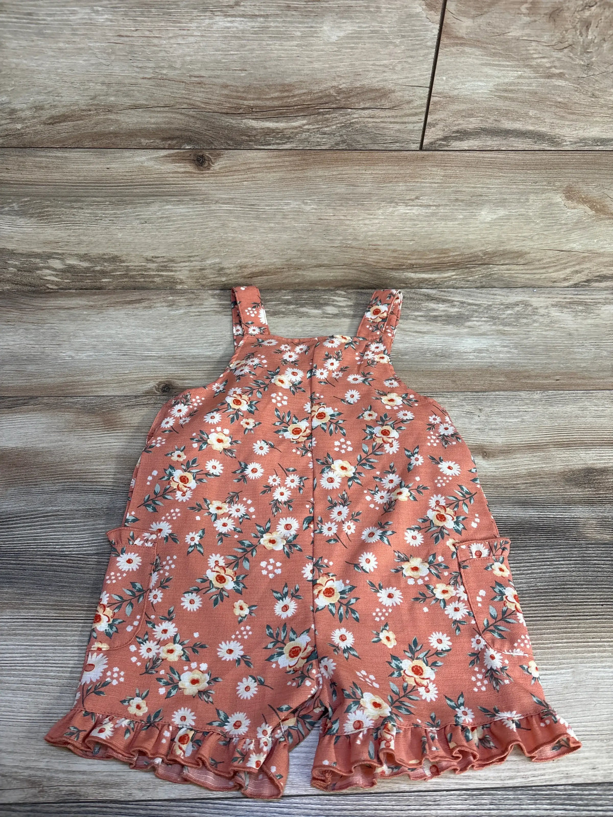 Flat lay of coral floral shortall (size 5T) showing ruffle hem, front straps, and side pockets on wood background