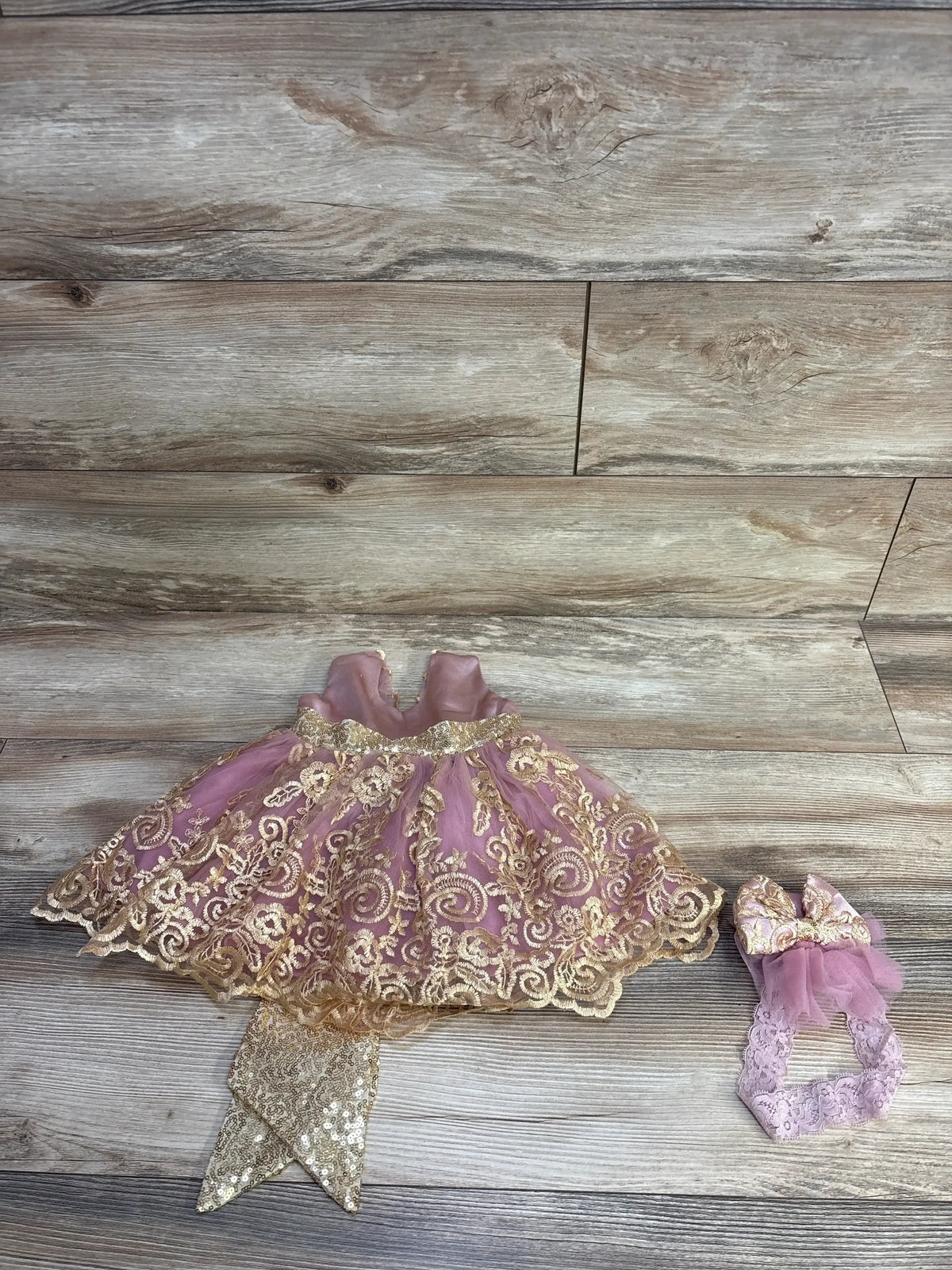 Flat lay of Melody 3-piece baby dress set, pink with gold lace and sequin sash, plus matching bow headband on wood background