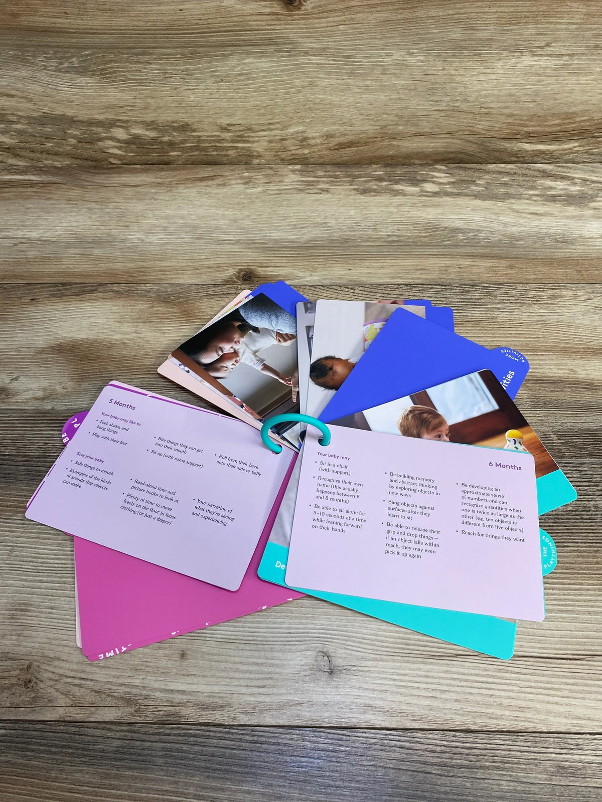 Angled view of Lovevery The Senser Play Guide flashcards showing 5- and 6-month milestone cards with photos and a teal binder ring on a wooden surface
