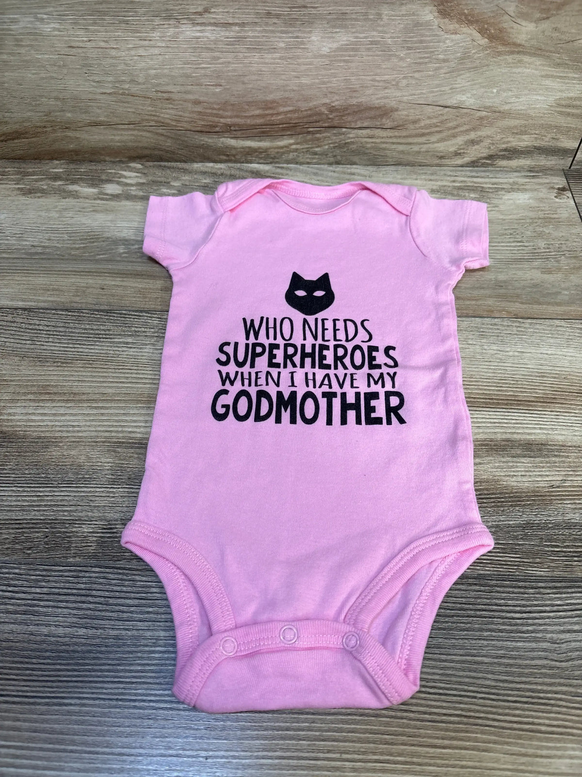 Top-down of pink Carter’s 12m bodysuit with black Who needs superheroes when I have my godmother text on wood background
