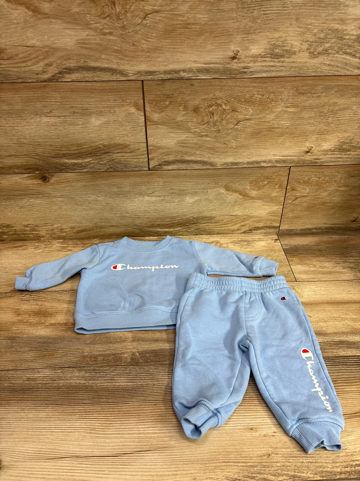 Front view of a blue Champion two-piece pullover sweatshirt and joggers set showcasing Champion logos on a wood background