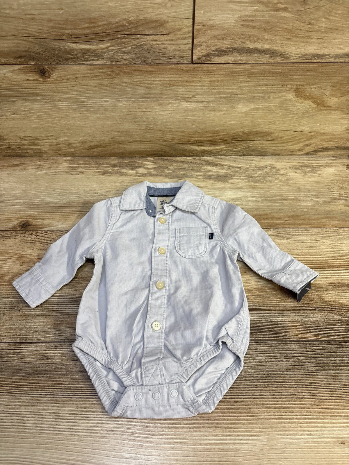 Flat lay Oshkosh corduroy baby bodysuit, light blue, button-up with chest pocket and snap crotch on wood background