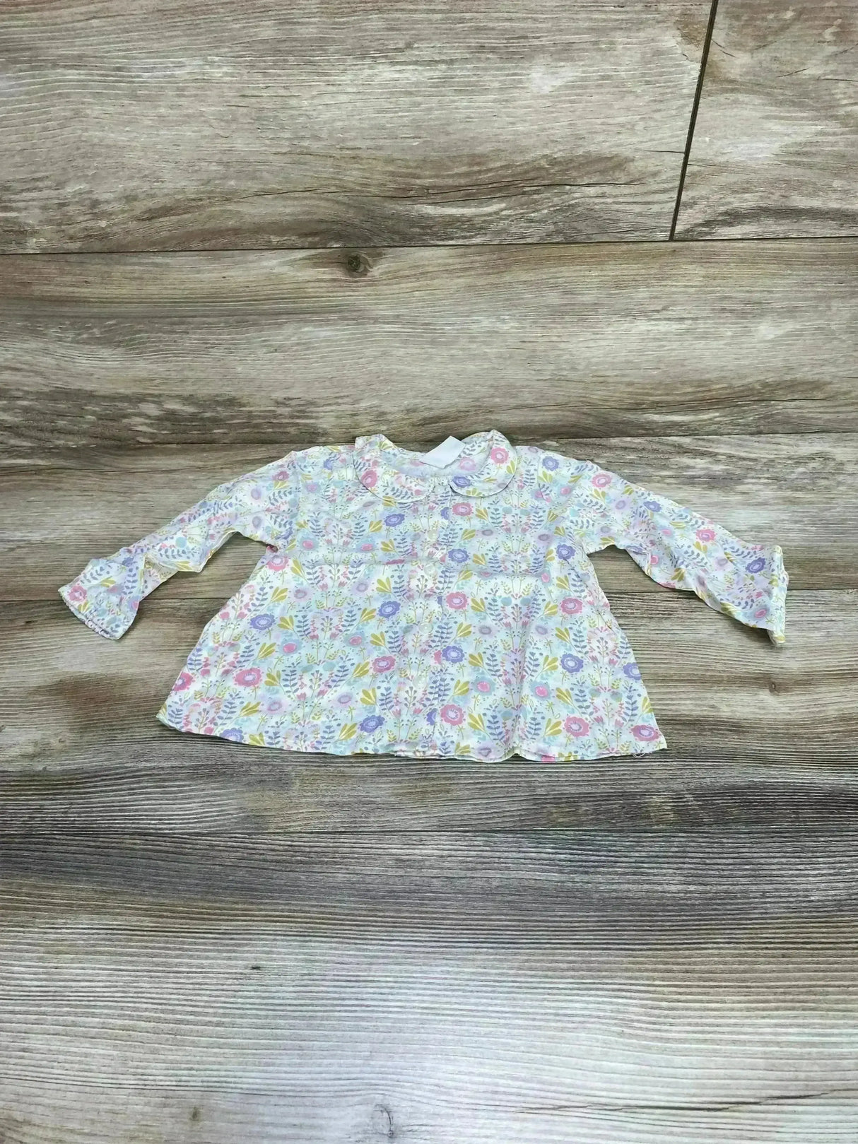 Flat lay of H&M baby top with pastel floral print, rounded collar, long sleeves, gathered waist on wood background