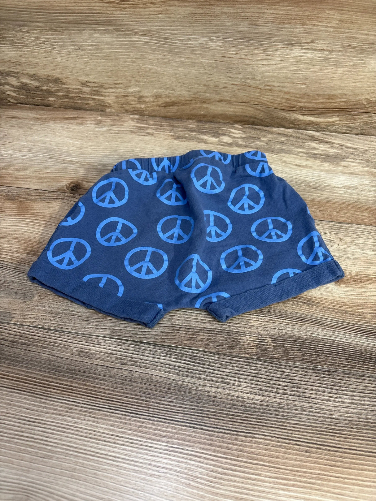 Front view of navy baby shorts with light blue peace sign print, elastic waistband and rolled hems on wooden surface