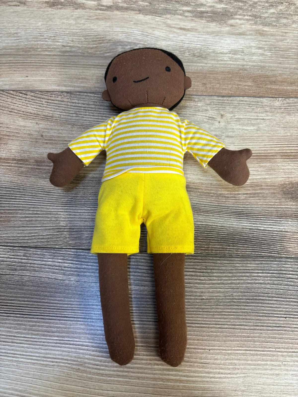 Front view of Cloud Island Plush Doll showcasing brown knit skin, yellow-and-white striped shirt and bright yellow shorts on wood plank background