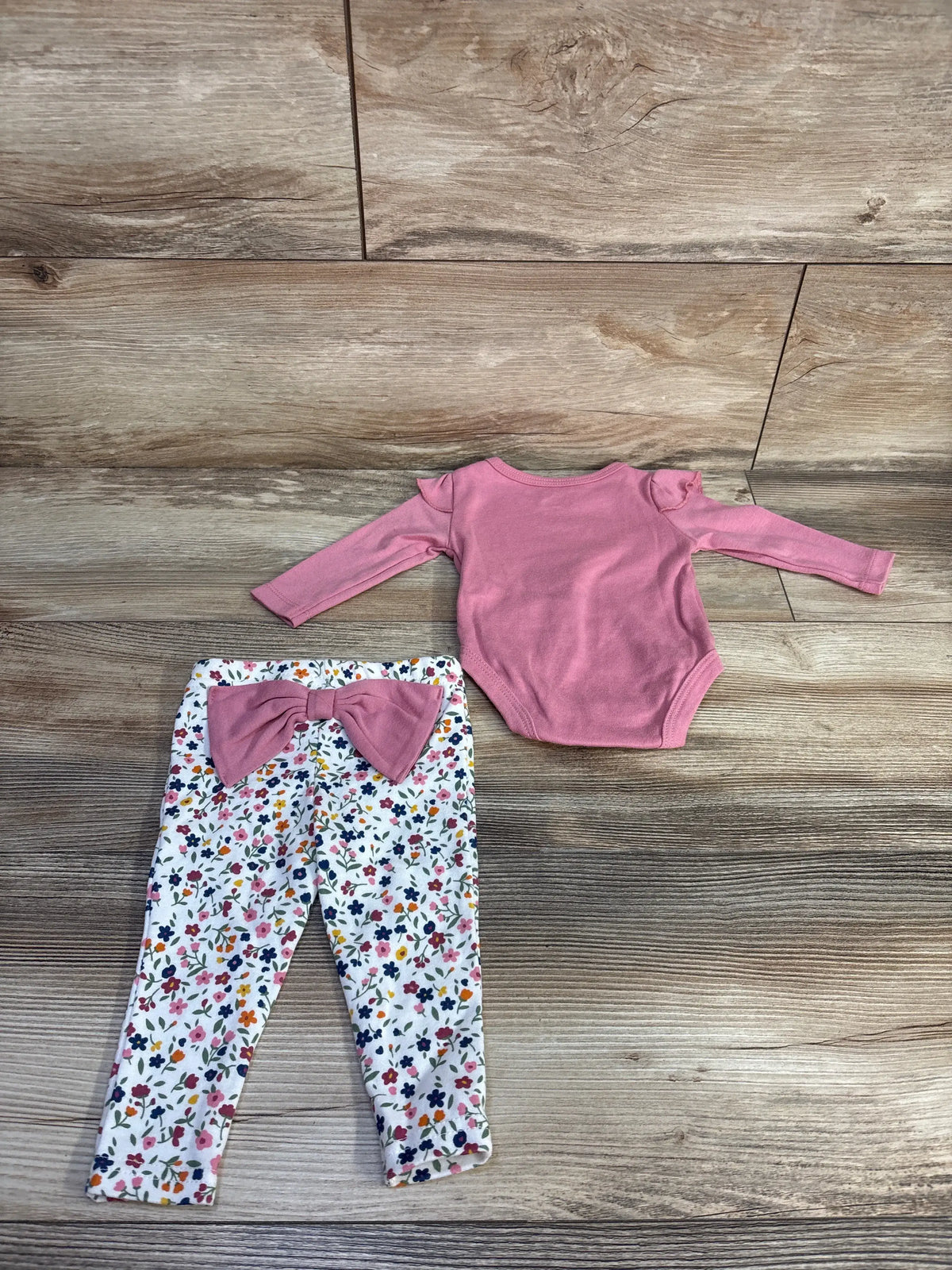 Top-down of 2-piece baby set: pink bodysuit with ruffled shoulders and white floral leggings with pink bow on wood